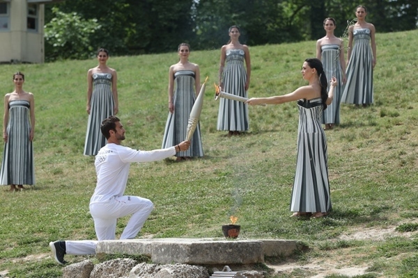 扮演最高女祭司的演員瑪麗·米娜（前右）點(diǎn)燃第一棒火炬手——希臘運(yùn)動(dòng)員、東京奧運(yùn)會(huì)男子賽艇項(xiàng)目金牌得主斯特凡諾斯·恩圖斯科斯手中的火炬。新華社記者 趙丁喆 攝