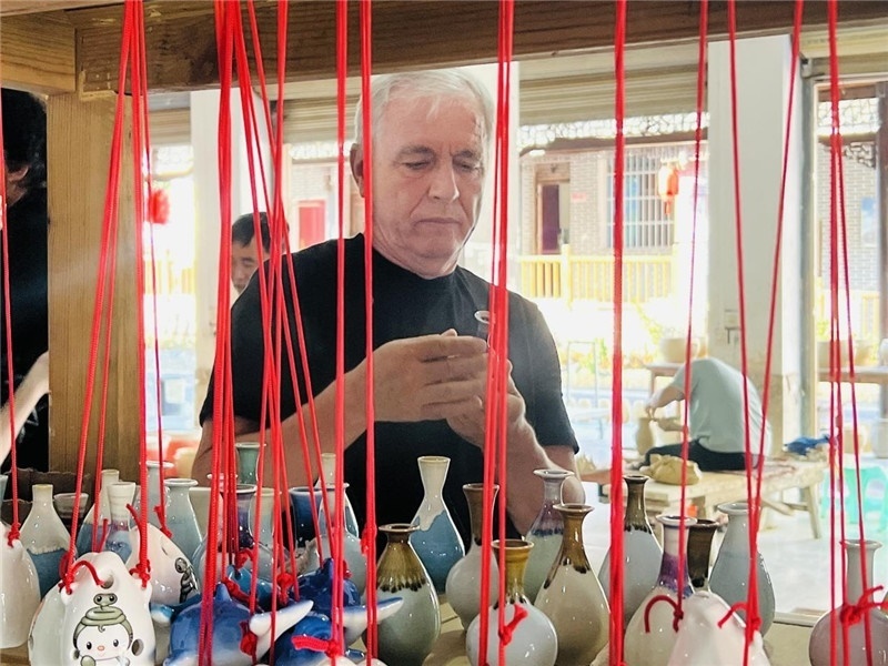 鄉(xiāng)愁如同一根根紅線，將游子與故鄉(xiāng)永遠(yuǎn)地牽連到一起?；逄账囄幕瘎?chuàng)意一條街的母氏陶藝館內(nèi)，紅線吊著陶瓶，思緒游走萬里。摩爾多瓦“信息標(biāo)簽”通訊社社長亞歷山大·塔納斯端詳著館內(nèi)展示的陶藝作品，并挑選出心儀的小擺件帶回摩爾多瓦，也帶走花茂村的“鄉(xiāng)愁”。（張亞欣攝）